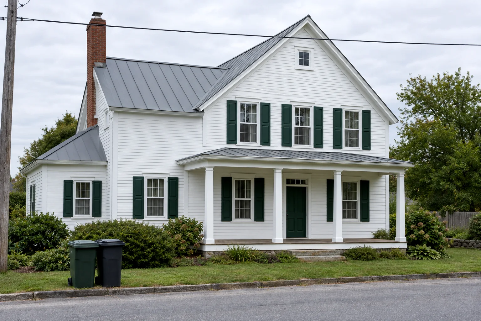 A white clapboard farmhouse with green shutters and a four-column porch, photographed from the street with two wheelie bins and a telephone wire in front