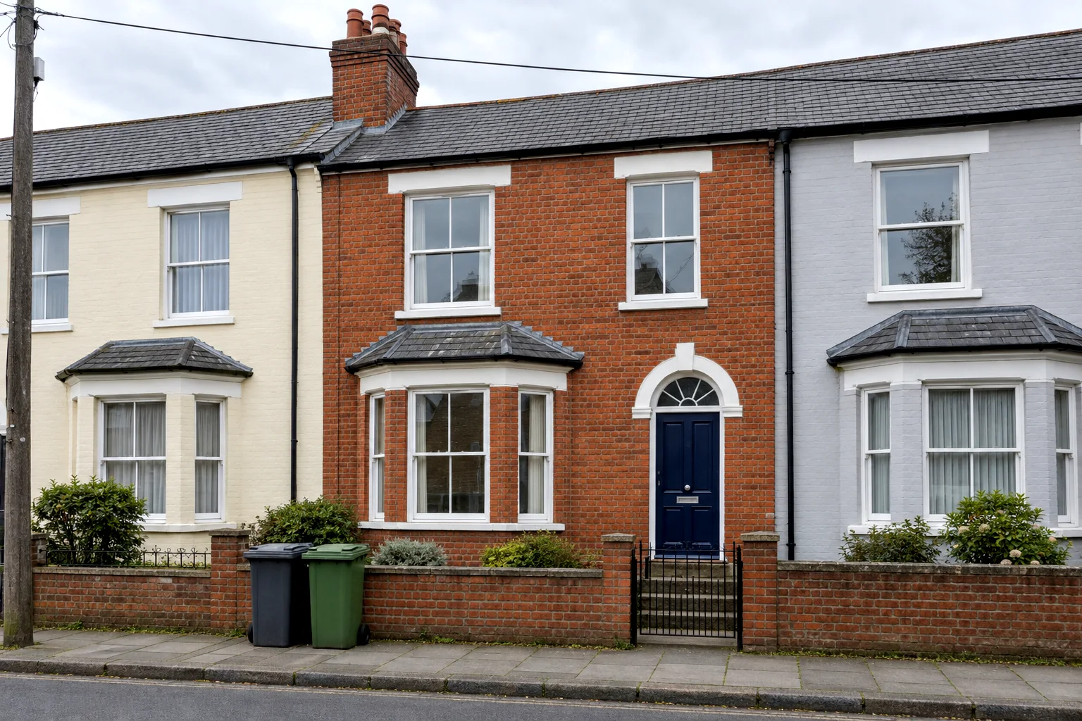 A red-brick terrace house with a bay window and a navy door between a cream and a grey neighbour, photographed from the street with two wheelie bins at the wall